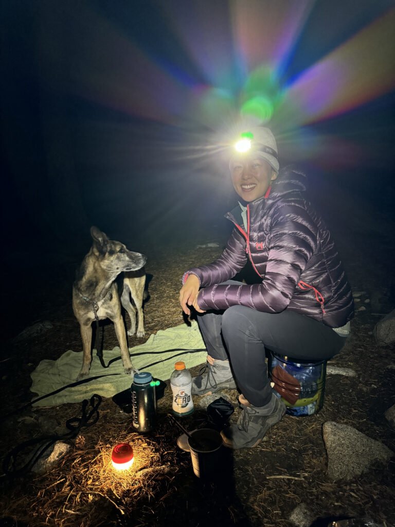 A camper seated, wearing a headlamp, with a dog to her left.